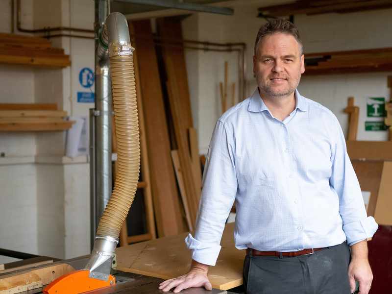 A teacher stands in a room, surrounded by woodworking equipment and tools.
