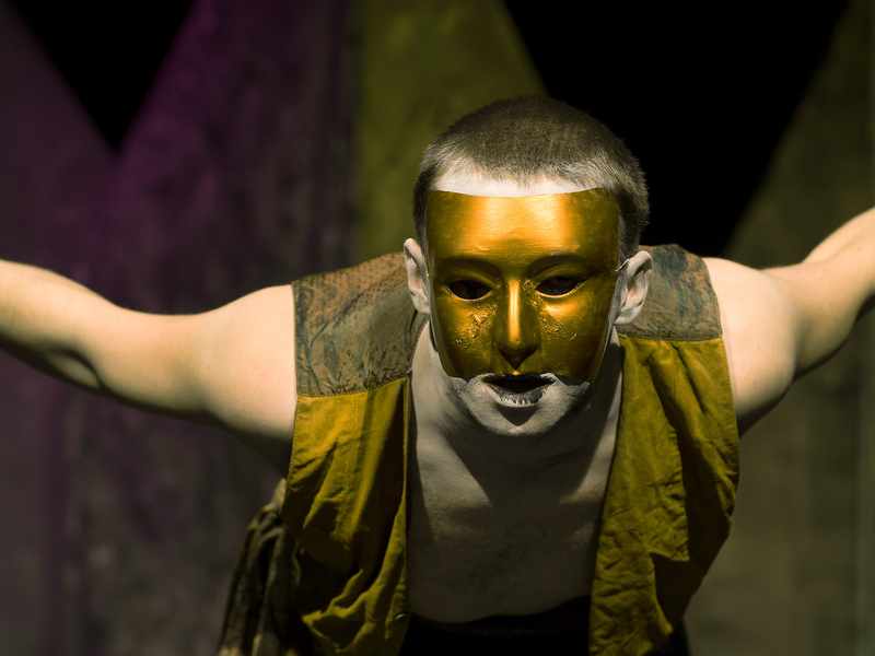 A performer leans forward with arms outstretched. He is wearing a gold mask covering his face.