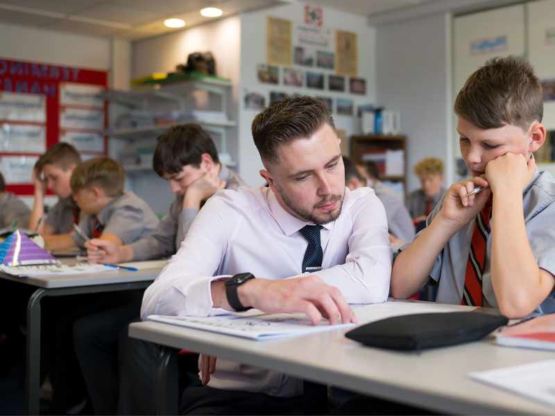 A teacher kneels next to a pupil sitting at a desk. He is pointing to the open page of a book.