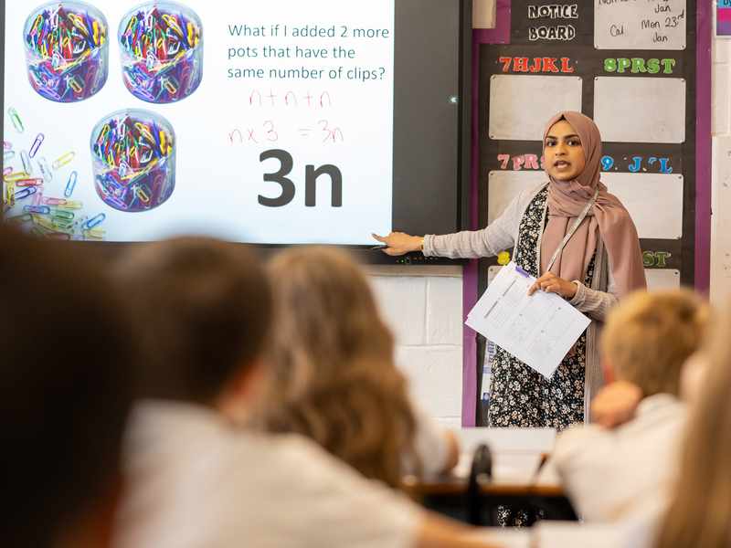 A teacher stands in front of a class. She is pointing to a whiteboard with algebra equations written on it.