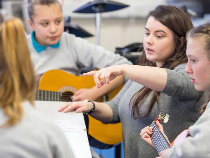 A teacher stands with a group of pupils. Two of the pupils are holding an acoustic guitar.