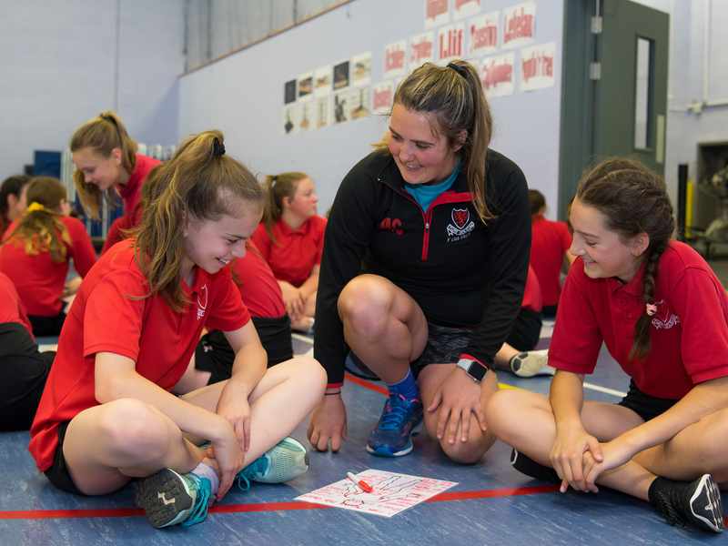 A teacher kneels between two pupils sitting on the floor of a gymnasium.