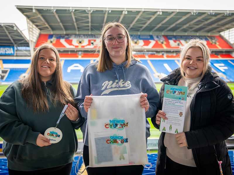 Three young women in the stands of a sports stadium hold Quick Change branded merchandise