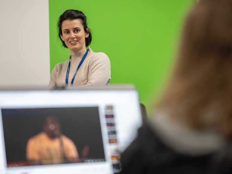 A person stands with their arms folded. Facing them is another person with an open laptop.