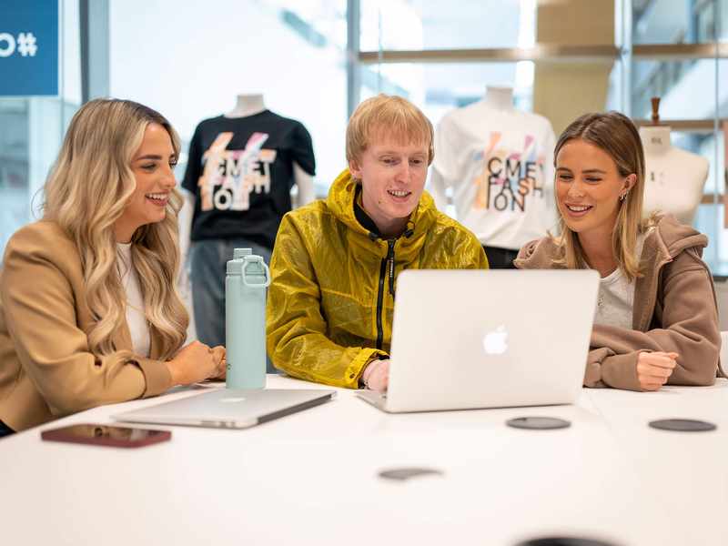 Three people sit alongside each other at a table with laptop computers.