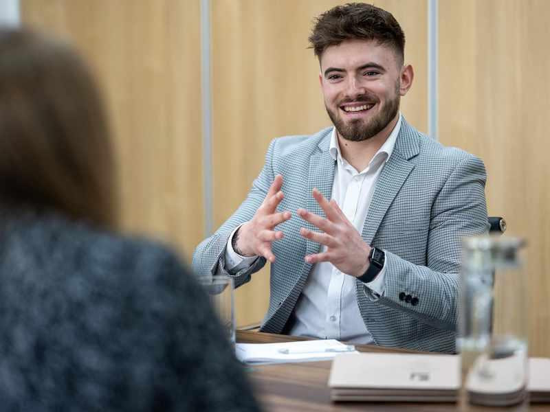 A person in a suit speaks to another person, both appearing engaged in a discussion.