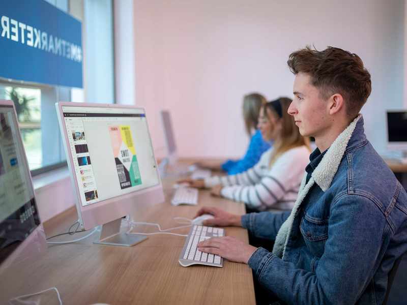 A person sat at a desk in front of a Mac computer. In the background are two other people also using computers.