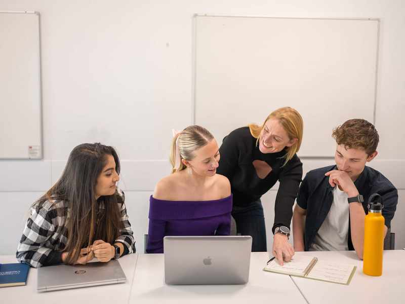 Three people sit alongside each other at a table with laptop computers and notebooks. Behind them another person leans in, pointing to an open notebook.
