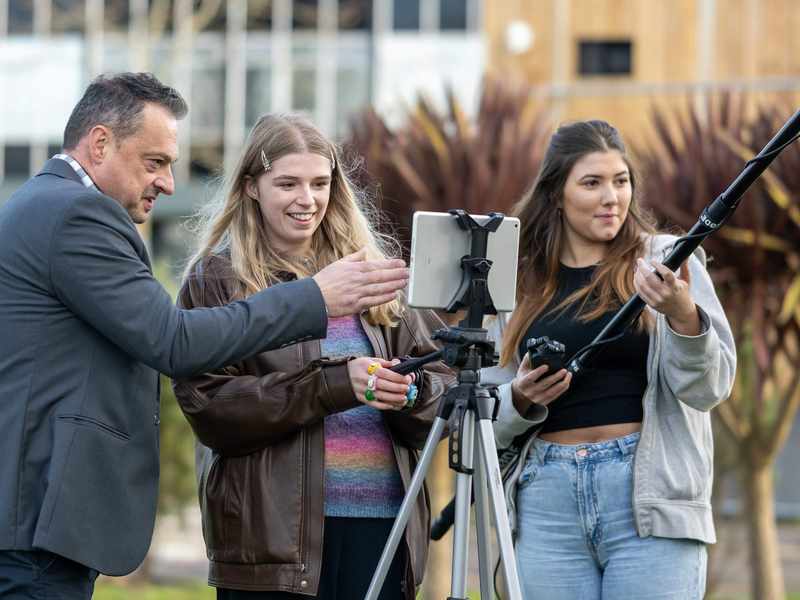 A group of people film outside. One person is operating a camera, while another holds a microphone boom.