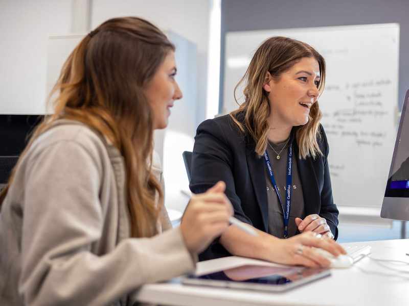 Two people sat at a desk in front of a Mac computer.