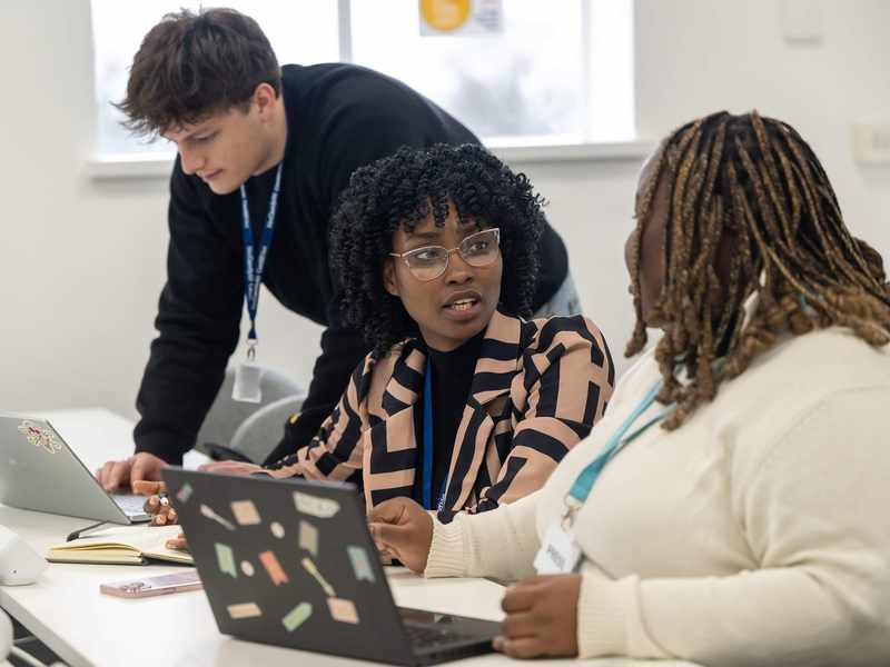 Three people working alongside each other at a desk. In front of them are two laptop computers.
