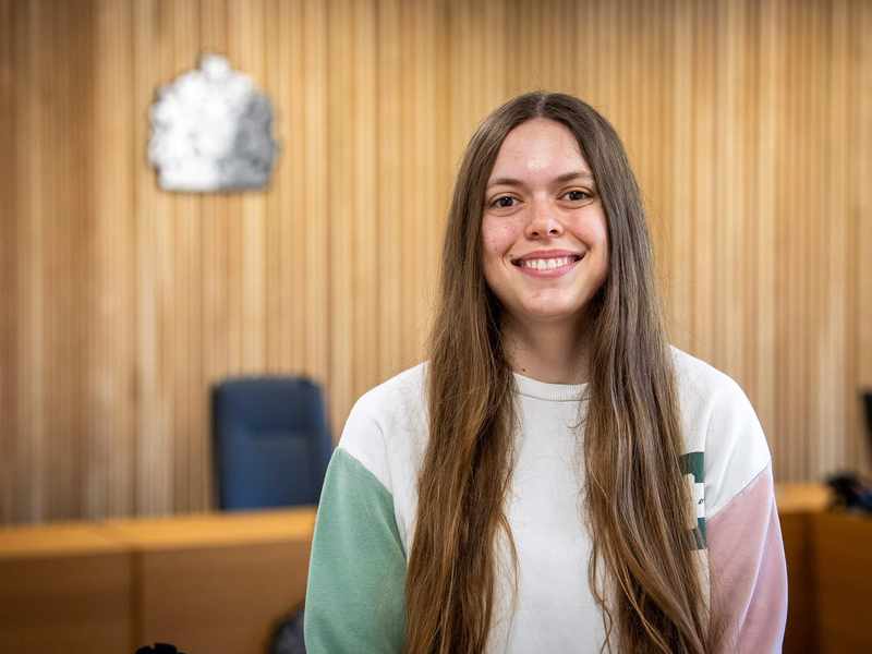 A person stands in a courtroom. In the background is a bench with a crown seal above it.