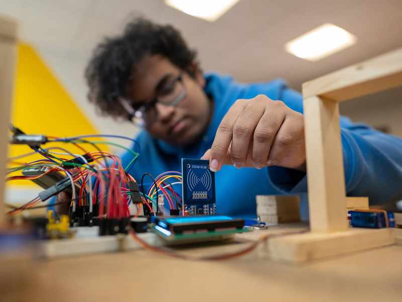 A person in a blue hoodie works with electronic components and circuit boards, placing a small blue board among connecting wires on a table. The background is blurred and the lighting is bright.