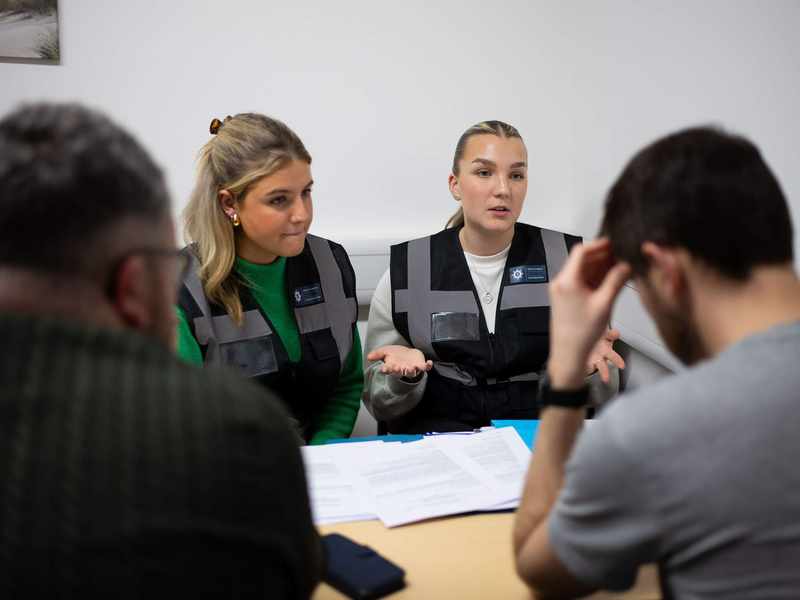 Two people wearing police vests sit at a desk facing another person.