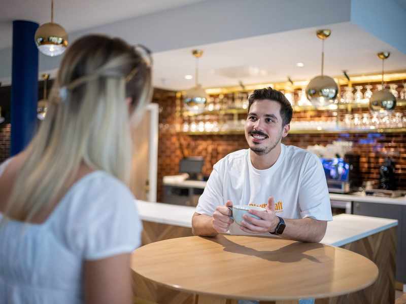 Two people sit at a table talking to each other. In the background is a bar area with a coffee machine.