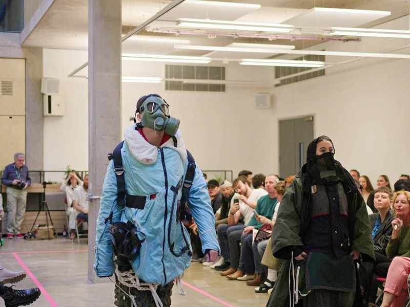 Two models walk along a fashion catwalk. They are wearing heavy coats and gas masks.