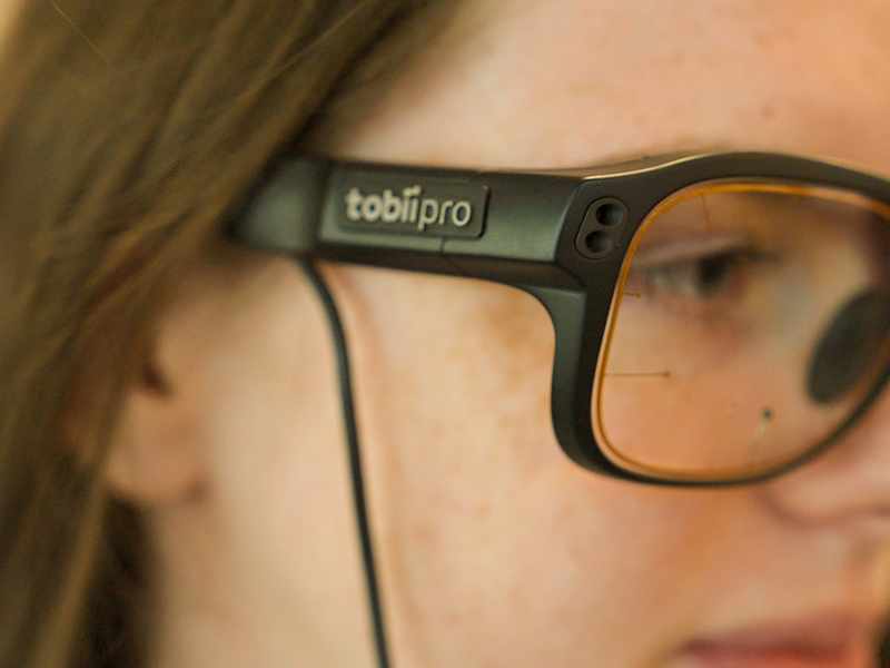 A close-up of a person wearing glasses. The glasses appear to have sensors on the front and a wire attached to the frame.