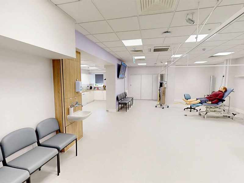 A large white room. In the background, a simulated patient lies on a trolley.