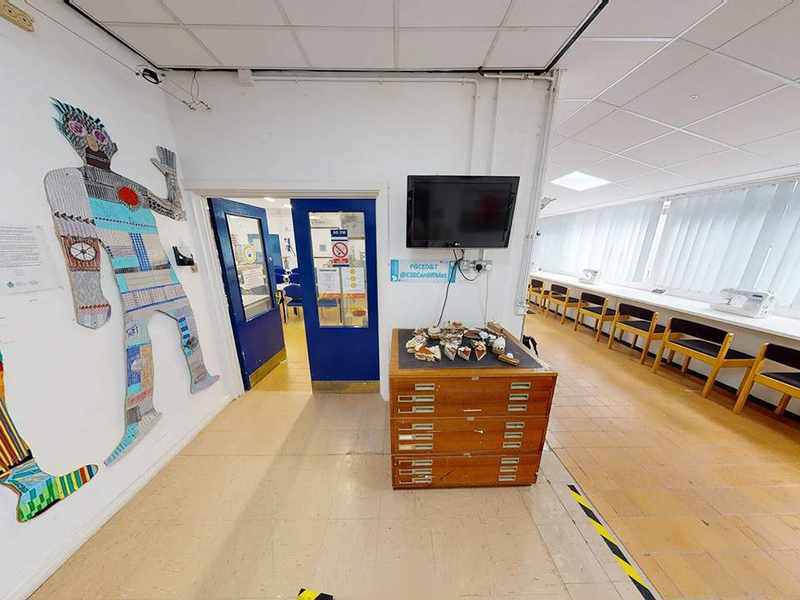 A corridor outside a room with desks and chairs. Mounted on the wall are stylised figures made from different materials.
