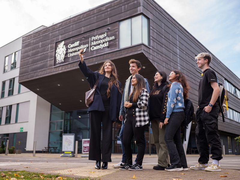 Six young adults stand huddled together outside the Cardiff School of Management as one of them takes a selfie on a mobile phone