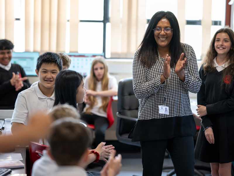 A teacher stands in a classroom surrounded by a group of pupils. There are computer monitors in the background.