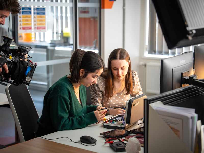 Two students work on a circuit board sat at a computer desk