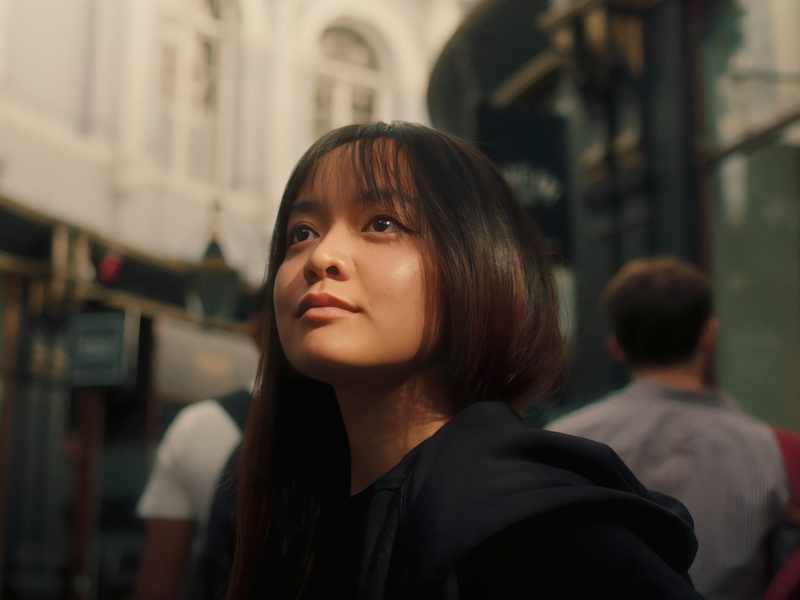 A woman gazes upward at the sky while strolling along a city street, embodying a moment of reflection and wonder.