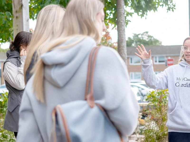 Young woman in grey hoodie and jeans gestures with hand while giving a campus tour to a group of visitors