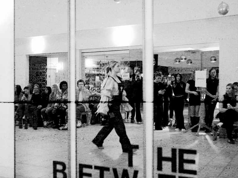 A black and white image of a mirror in which the reflection of a catwalk fashion show with attendees can be seen