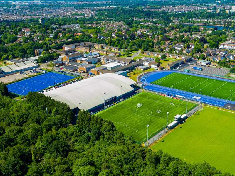 Aerial view of a sports complex featuring a large indoor sports facility, blue athletics track, multiple soccer fields, lush green trees in the foreground, and nearby residential buildings in the background.