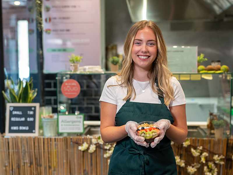 A young person wearing a green apron holds a fruit bowl while standing in front of a kitchen counter