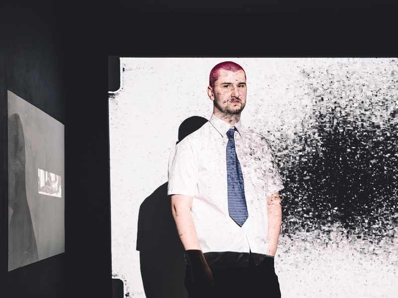 A young man in white shirt and tie stands in the light of a projector screen