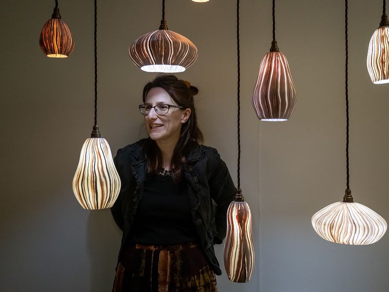 A woman wearing glasses leans against a white wall behind a collection of hanging lights