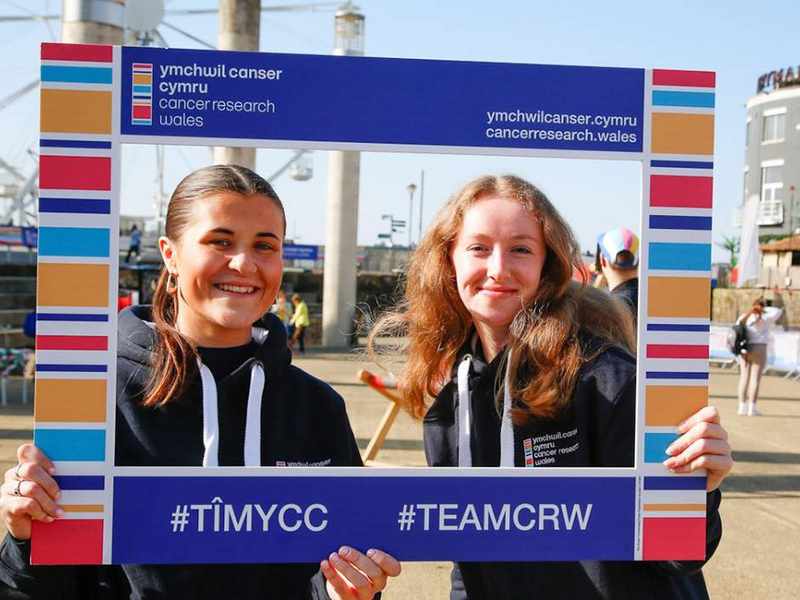 Two people hold a sign for Cancer Research Wales.