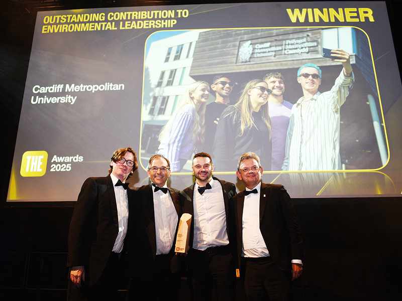Cardiff Met staff accept an environmental leadership award on stage beneath a winner announcement screen.