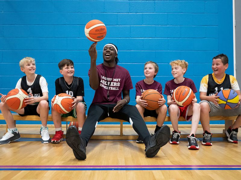 A sports coach spins a basketball on his fingers while four pupils, all holding a basketball on their laps, watch on