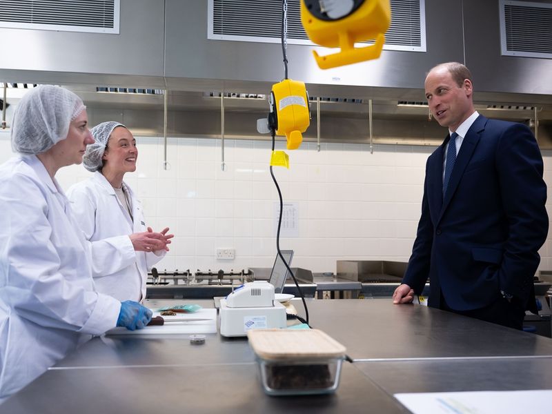 Prince William engages in conversation with two workers in lab coats and hairnets