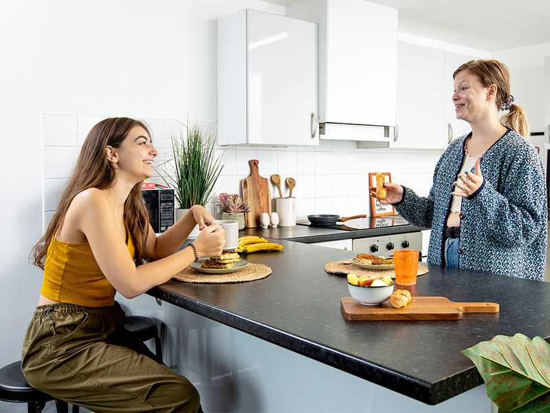 Two female students engaged in conversation while sitting at a kitchen counter, surrounded by kitchen items and warm lighting.