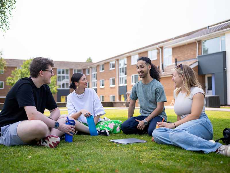 Group of four students sitting on the grass, chatting and laughing outside a student accommodation building