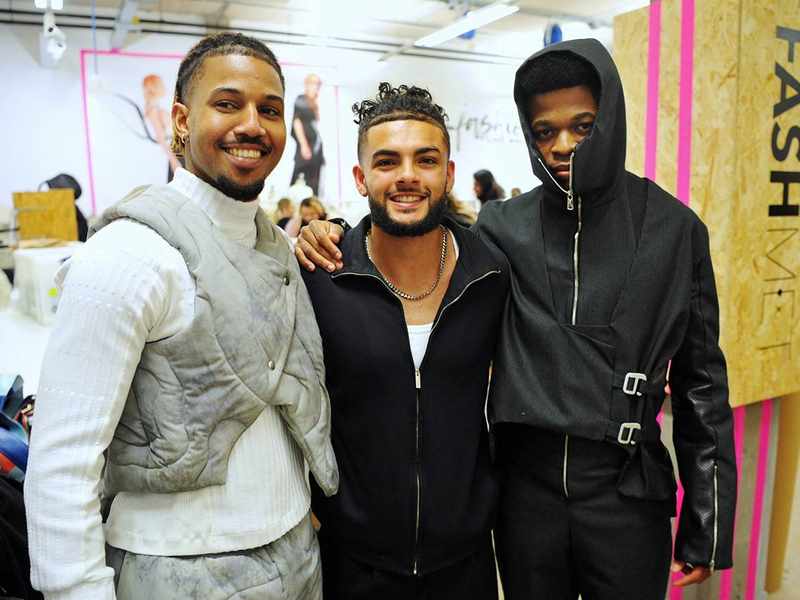 Three people pose arm in arm. In the background, costume sketches can been seen on the wall.