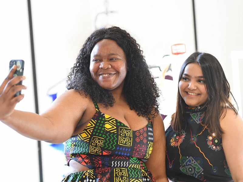 Two people pose for a selfie. They are wearing black dresses, patterned with bright colours.