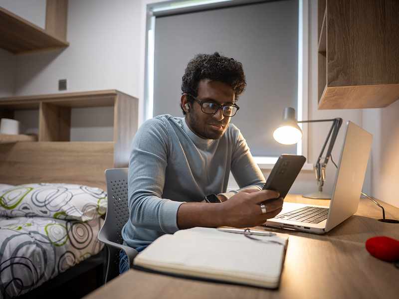 A man seated at a desk, working on a laptop while holding a cell phone in his hand.