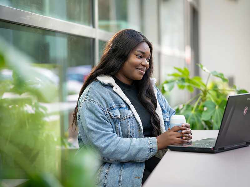 A young woman sits at an outdoor table holding a coffee in both hands while reading from their laptop screen.