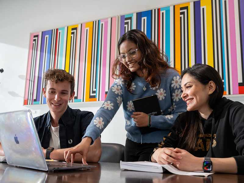 Three students collaborating on a laptop in a modern office setting, focused on their project.