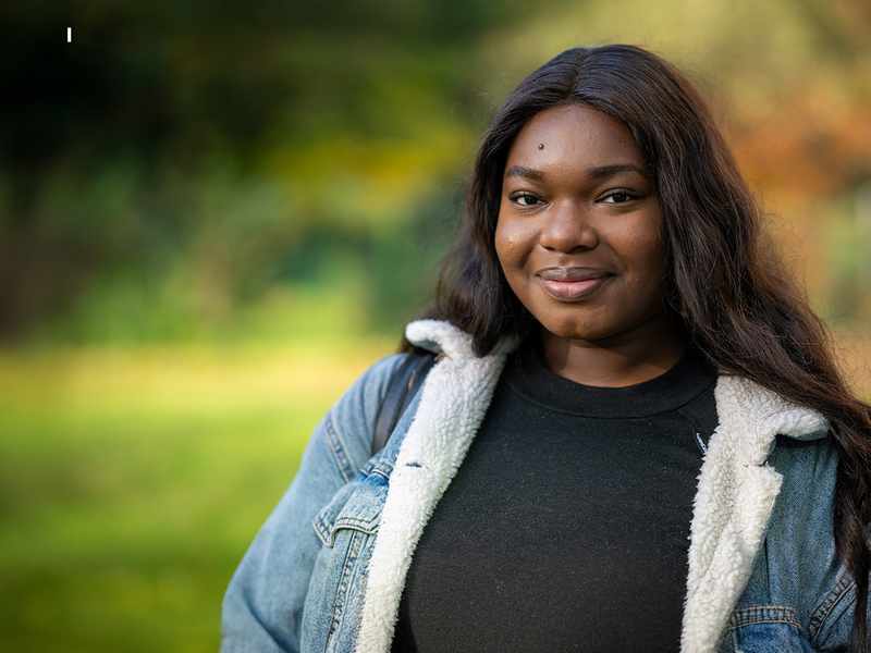 A young woman wearing a black shirt and a denim jacket, smiling confidently against a neutral background.