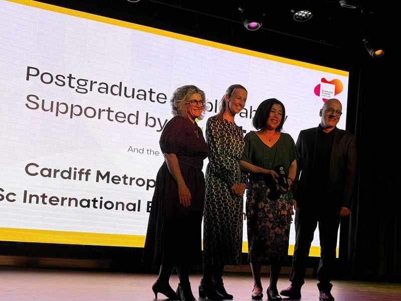A group of four people pose for a photo in front of large display screen. One of them is holding an award in their hands.