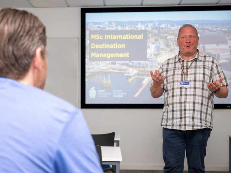 A man stands in front of a screen giving a presentation. The screen shows a view of Cardiff Bay with the caption MSc International Destination Management.