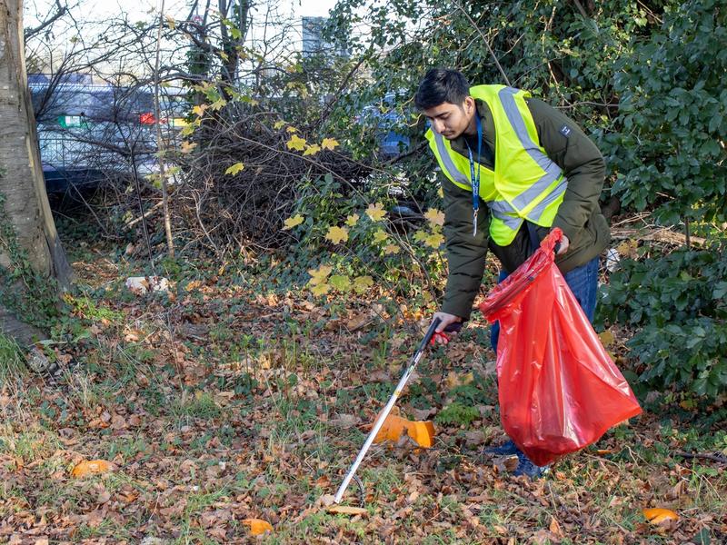 Young adult in high visibility jacket uses a litter picker to collect rubbish in a large red bin bag
