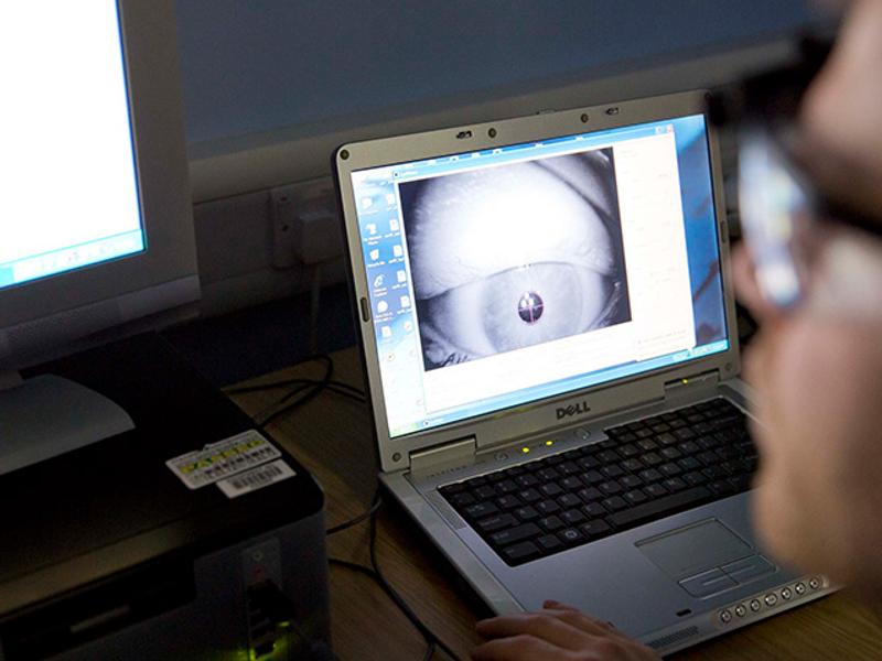 A close-up of a person working on a Dell laptop displaying an eye-tracking software interface. The screen shows a magnified image of an eye being analysed. A second computer monitor and other lab equipment are visible in the background.