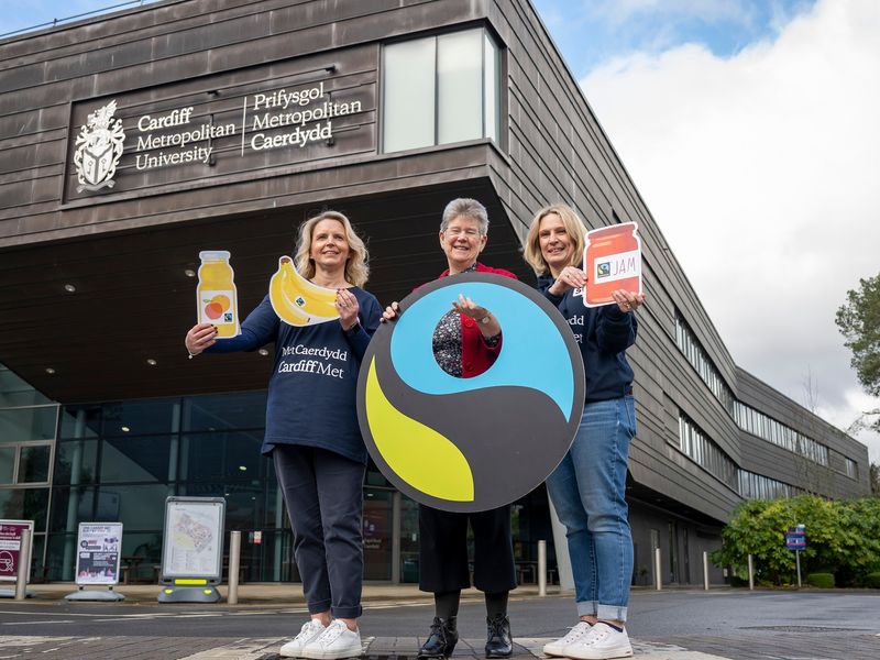 Three members of Cardiff Met staff hold Fairtrade information posters outside the Cardiff School of Managament